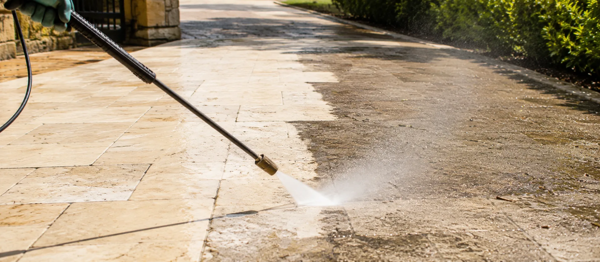 A pressure washer lance stripping grime from limestone paving, leaving a clean line across the driveway